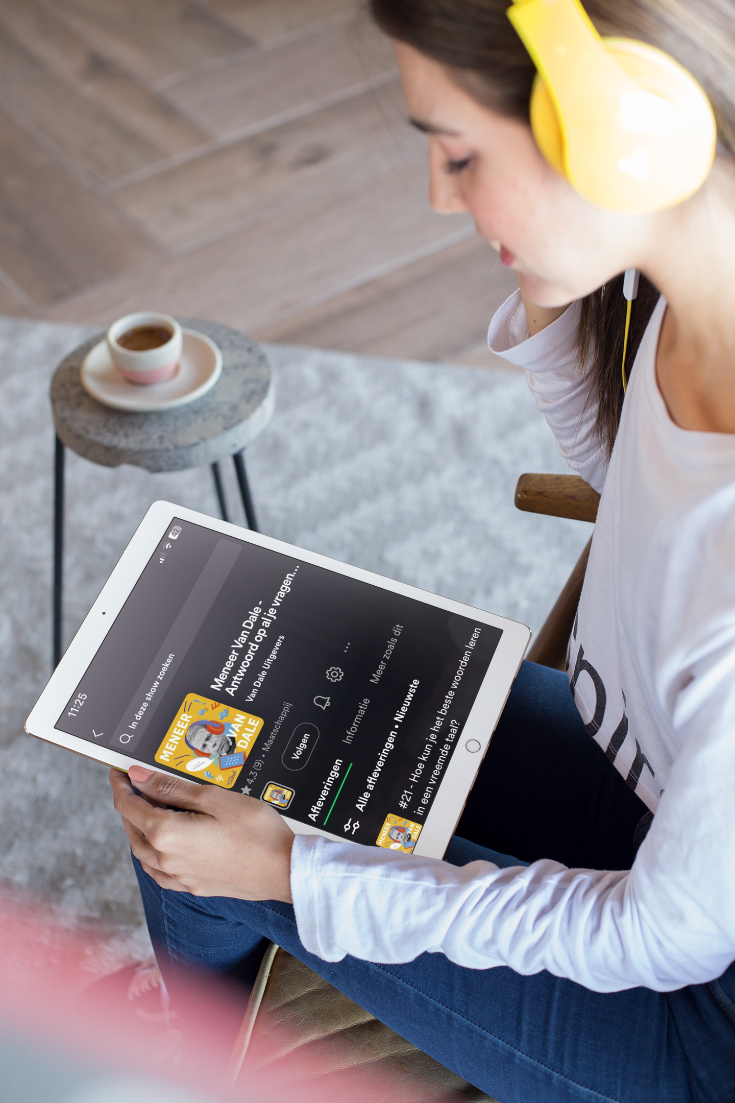 Een persoon heeft een koptelefoon op en een tablet op schoot. Op het tabletscherm is de Meneer Van Dale-podcast te zien