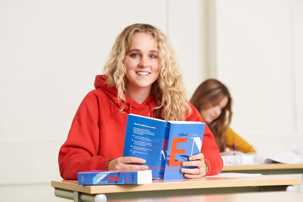 Twee scholieren zitten aan schooltafels. Een scholier zit in volle focus in het midden van de afbeelding. Ze kijkt naar de camera en heeft het 'Van Dale Pocketwoordenboek Engels-Nederlands' in haar handen. De andere scholier is niet in focus en is over diens tafel gebogen met een pen in diens hand.