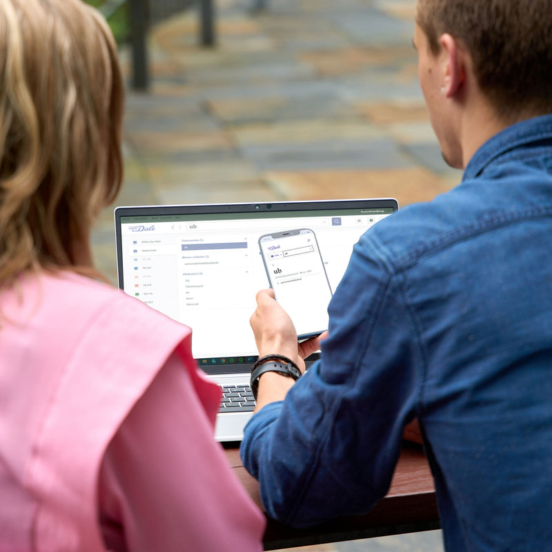 Twee studenten zitten met hun rug naar de camera toe. Tussen hen in staat een laptop met een schermafbeelding van Van Dale online. De rechter student heeft een telefoon vast die hij voor het laptopscherm houdt. Daarop is ook een schermafbeelding van Van Dale Online te zien. Op beide schermafbeeldingen staat de definitie van het woord 'ub'
