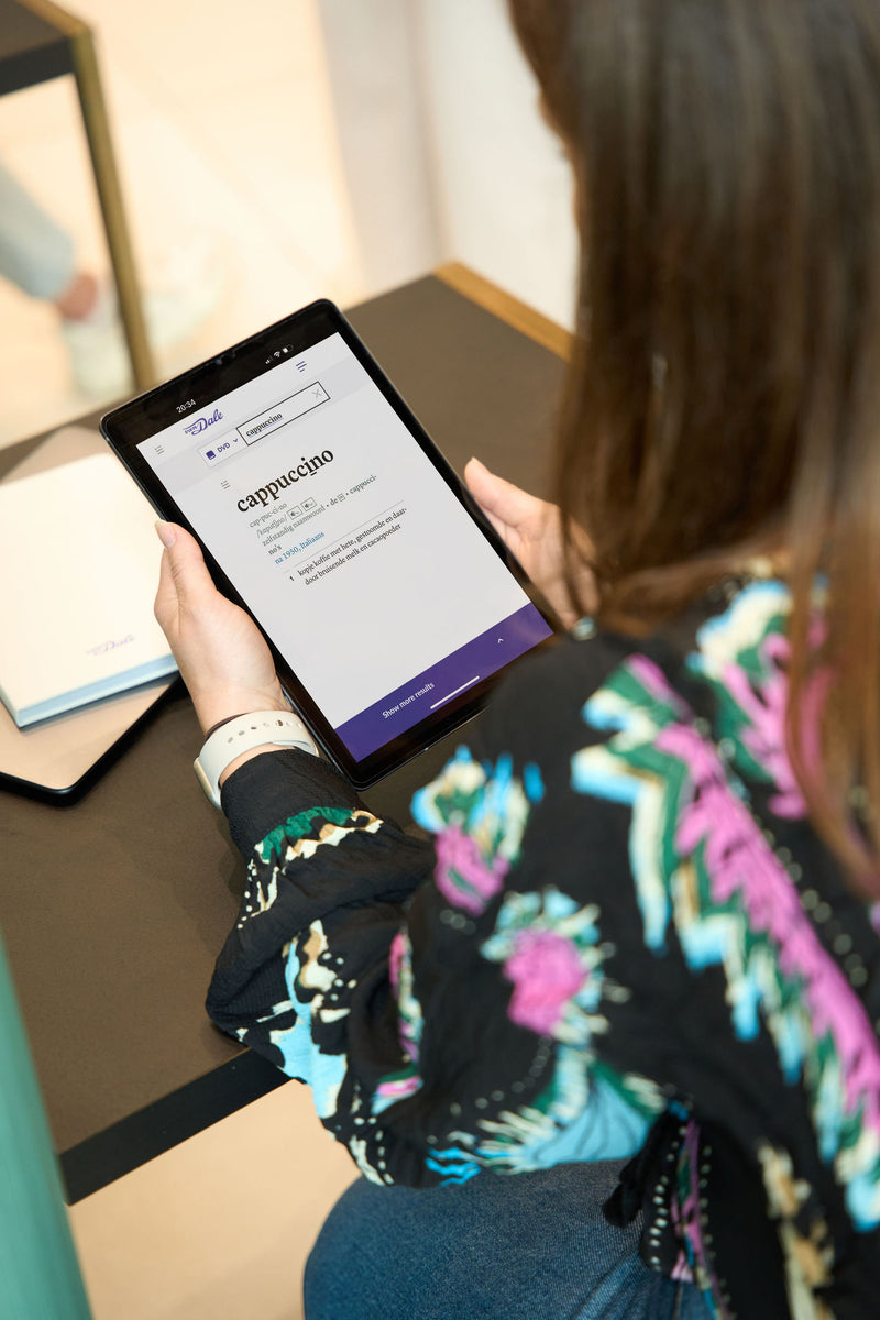 Een student is over een tablet gebogen met een schermafbeelding van Van Dale Online met de definitie van het woord 'cappuccino'