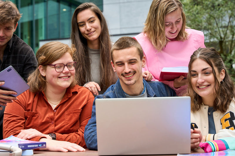 Vijf studenten, waarvan vier kijken naar de laptop die op tafel staat. Een van de studenten kijkt in een opengeslagen boek in diens handen.